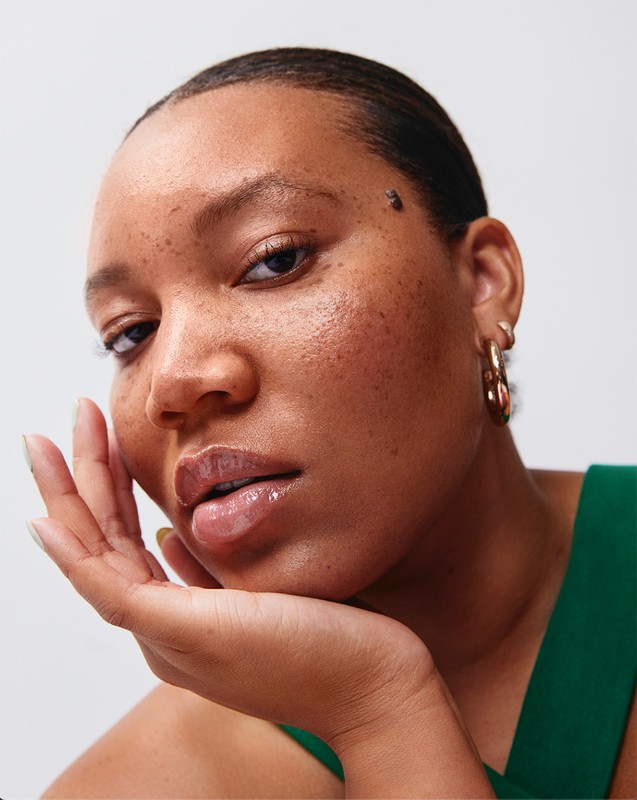 Close-up portrait of an adult woman with brown skin, dark eyes, and gold hoop earrings posing against a neutral white backdrop.