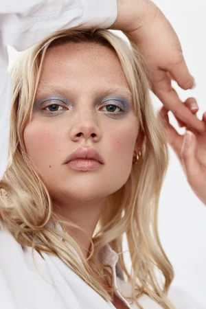 Close-up portrait of a blonde woman with metallic blue eyeshadow and freckles wearing a white shirt against a plain white background