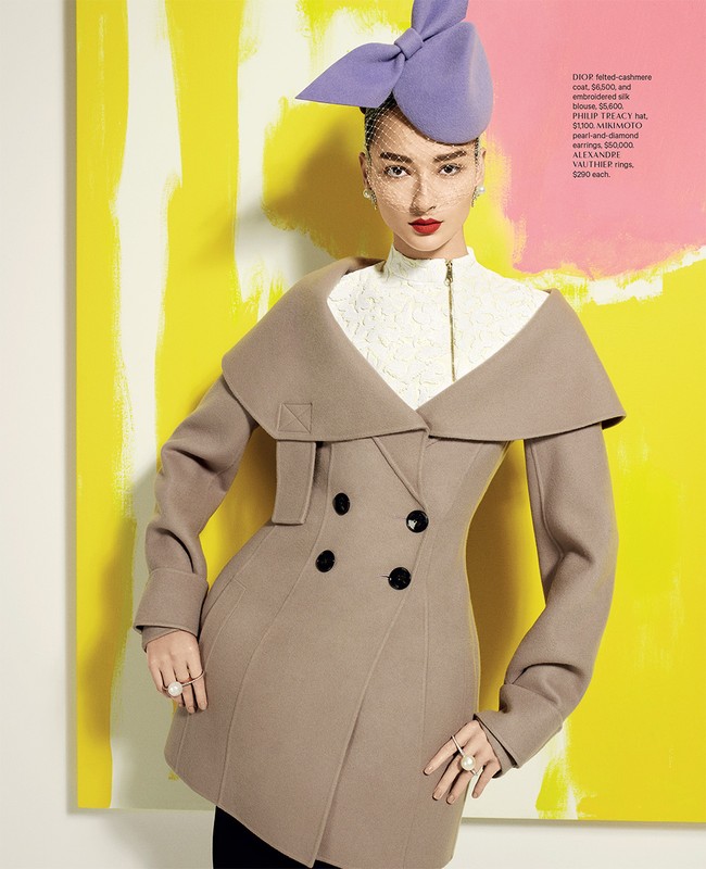 A fashion model poses in a beige double-breasted coat, white top, and purple hat against a yellow and white painted backdrop, exuding high fashion.