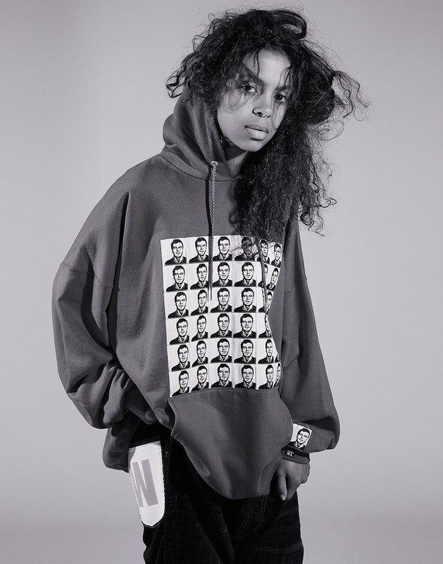Young woman in a hoodie with black-and-white graphics stands indoors against a plain backdrop in a studio portrait.