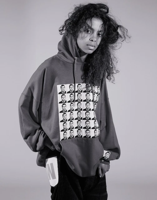 Young woman in a hoodie with black-and-white graphics stands indoors against a plain backdrop in a studio portrait.