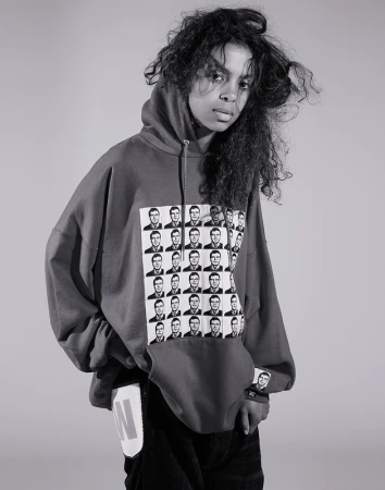 Young woman in a hoodie with black-and-white graphics stands indoors against a plain backdrop in a studio portrait.