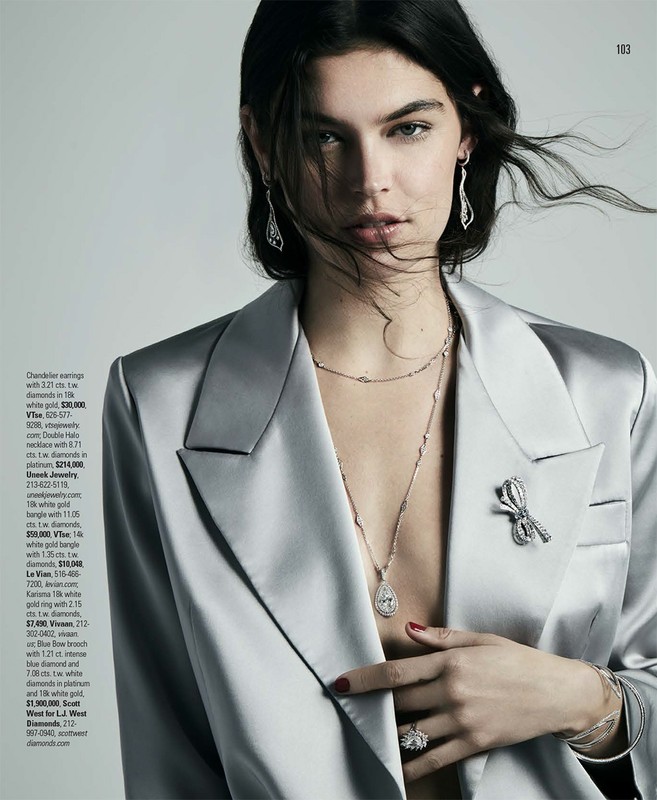 Close-up portrait of a woman wearing a glossy silver blazer, diamond pendant necklace, drop earrings, and a bow brooch against a light gray background.