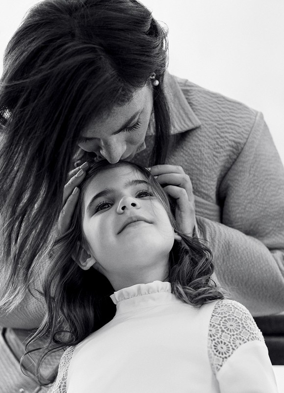 Monochrome portrait of a dark-haired adult female kissing the forehead of a girl in a white top. The image is eye-level, indoor, black and white.