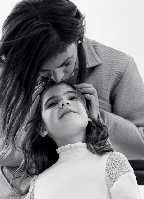 Monochrome portrait of a dark-haired adult female kissing the forehead of a girl in a white top. The image is eye-level, indoor, black and white.