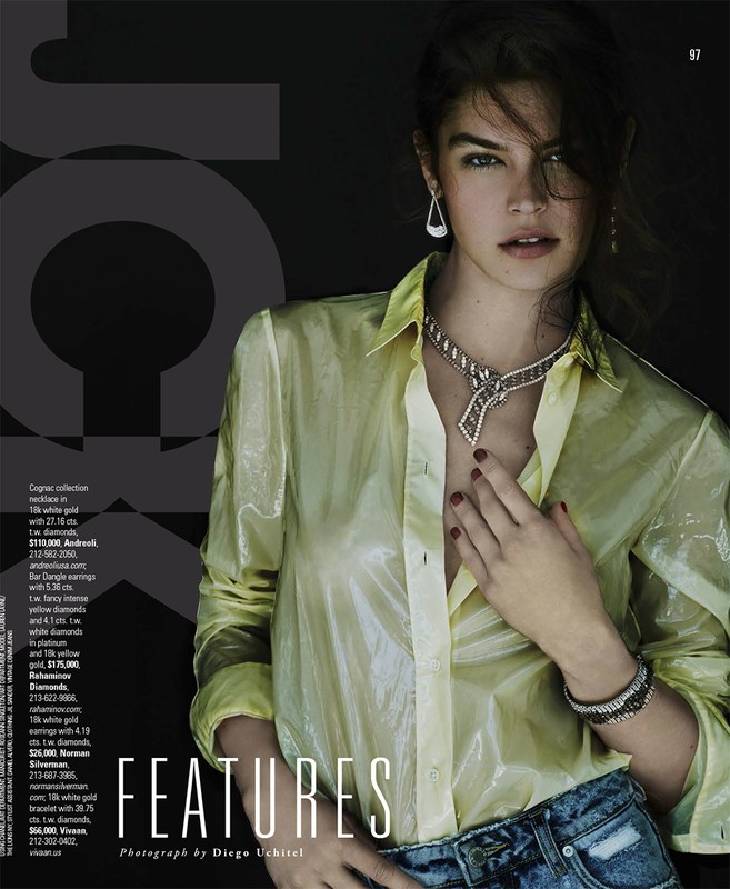 Editorial fashion shot of a woman wearing a sheer yellow blouse and jewelry including a necklace, earrings, and bracelet in a dark indoor space.