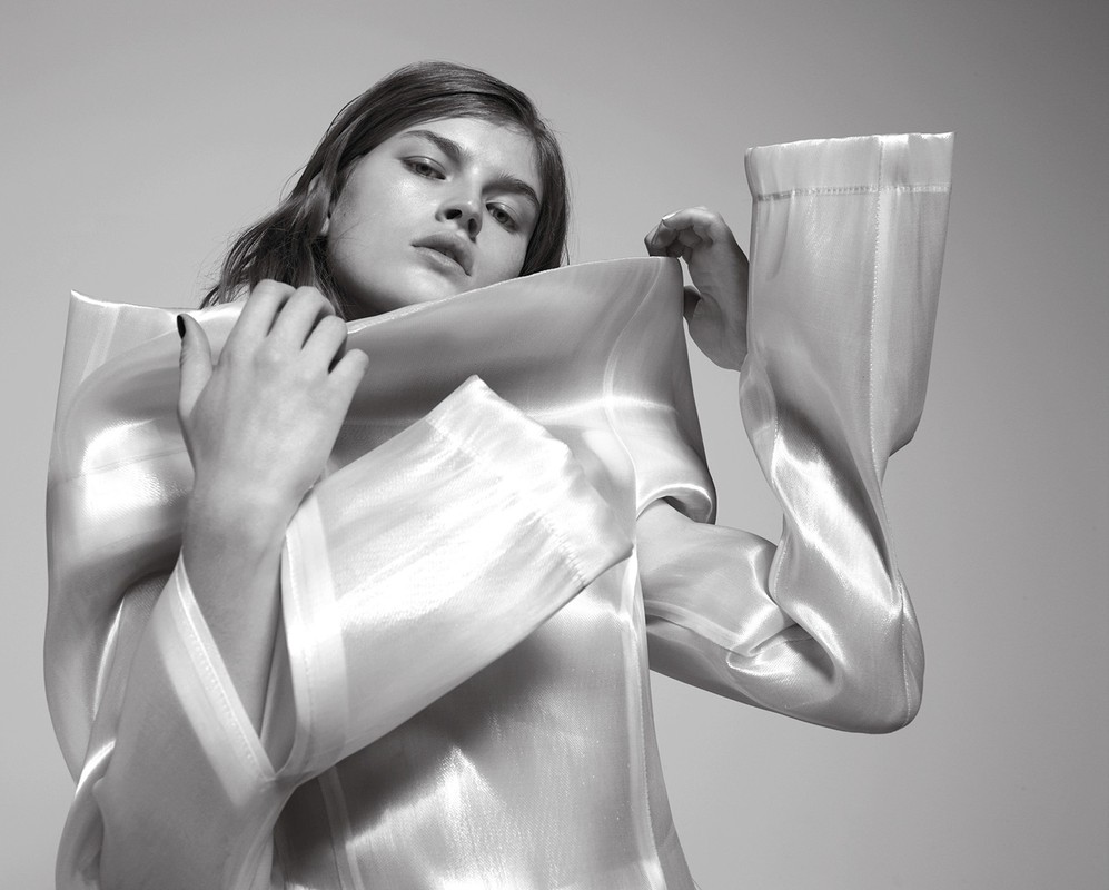 Elegant portrait of a woman in a flowing, sheer top, hands near her face, set against a neutral background in a fashion style.