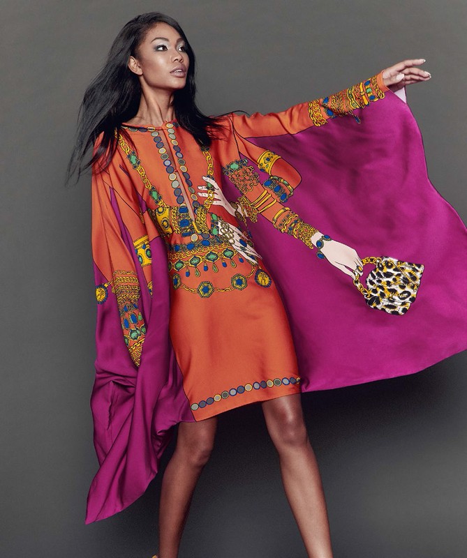 Fashion shot of a woman in an orange and purple dress with jewelry designs, holding a leopard print bag against a grey backdrop.