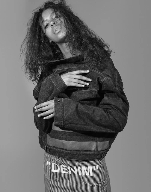 Black and white portrait of a stylish African American woman wearing a denim jacket and striped pants with the word "DENIM" printed on them.