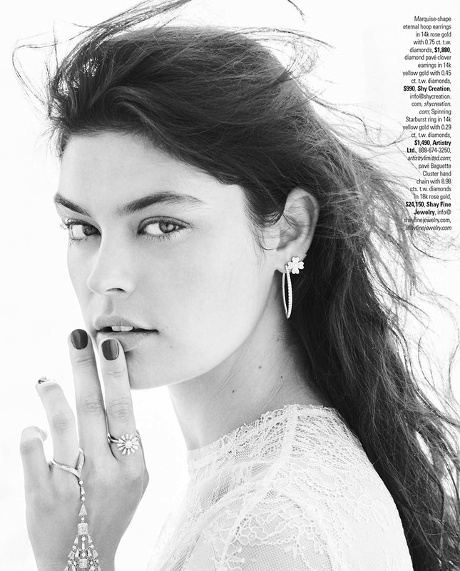Black and white close-up of a woman with dark hair wearing hoop earrings and rings, posing with fingers near her lips in a studio.