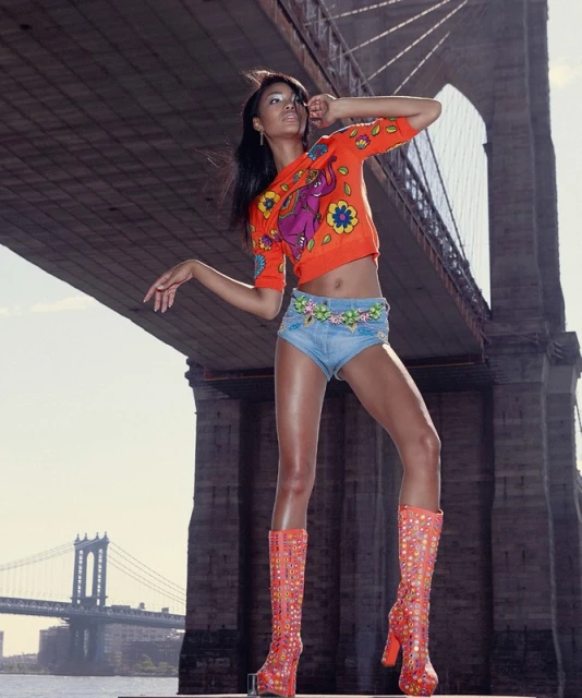 Fashion shot of a female model in a printed orange top, denim shorts, and tall boots, posing in front of the Brooklyn Bridge.