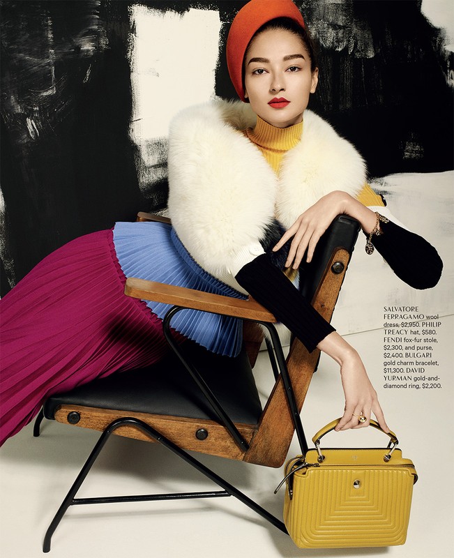 Woman in orange hat and yellow turtleneck poses in chair with handbag and pleated skirt in a fashion editorial against abstract background.