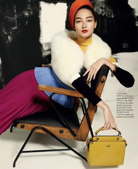 Woman in orange hat and yellow turtleneck poses in chair with handbag and pleated skirt in a fashion editorial against abstract background.
