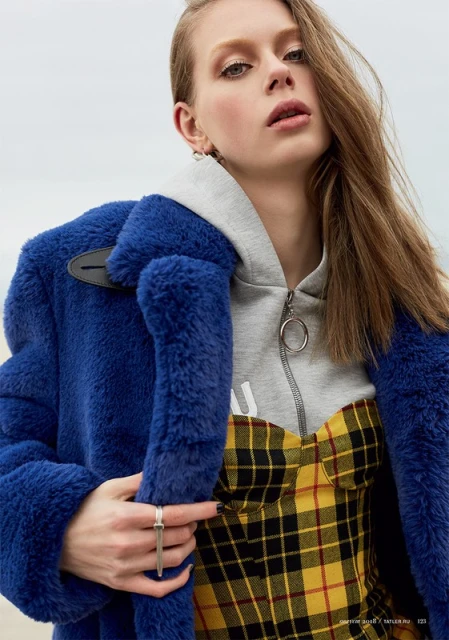 Close-up portrait of a fair-skinned woman with long brown hair, wearing a blue fuzzy coat and a yellow plaid dress, posing indoors.