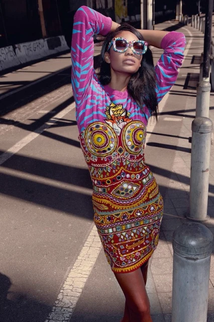 Model poses on city street wearing a colorful patterned mini dress and sunglasses in bright sunlight, long shot.