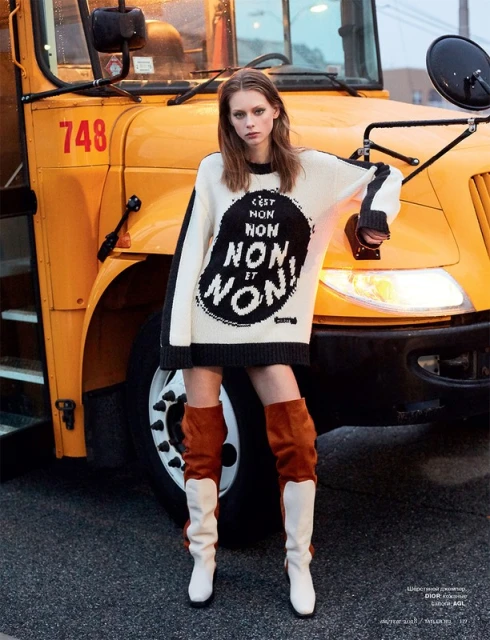 A young woman poses in a long sweater and high boots in front of a yellow school bus for a fashion editorial shot.