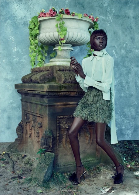 Elegant fashion portrait of a woman in a stylish green skirt and white blouse, posing next to a large stone flower pot with pink flowers.
