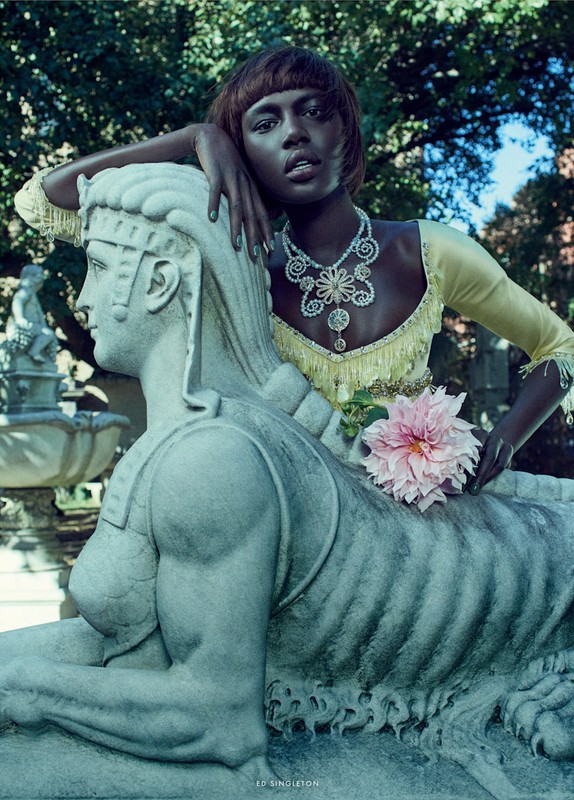 Fashion shot of African American woman in yellow dress and jewelry, posing with a stone sphinx statue in outdoor garden setting.