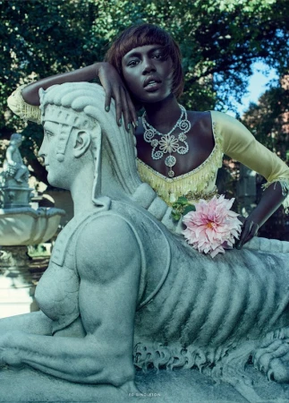 Fashion shot of African American woman in yellow dress and jewelry, posing with a stone sphinx statue in outdoor garden setting.