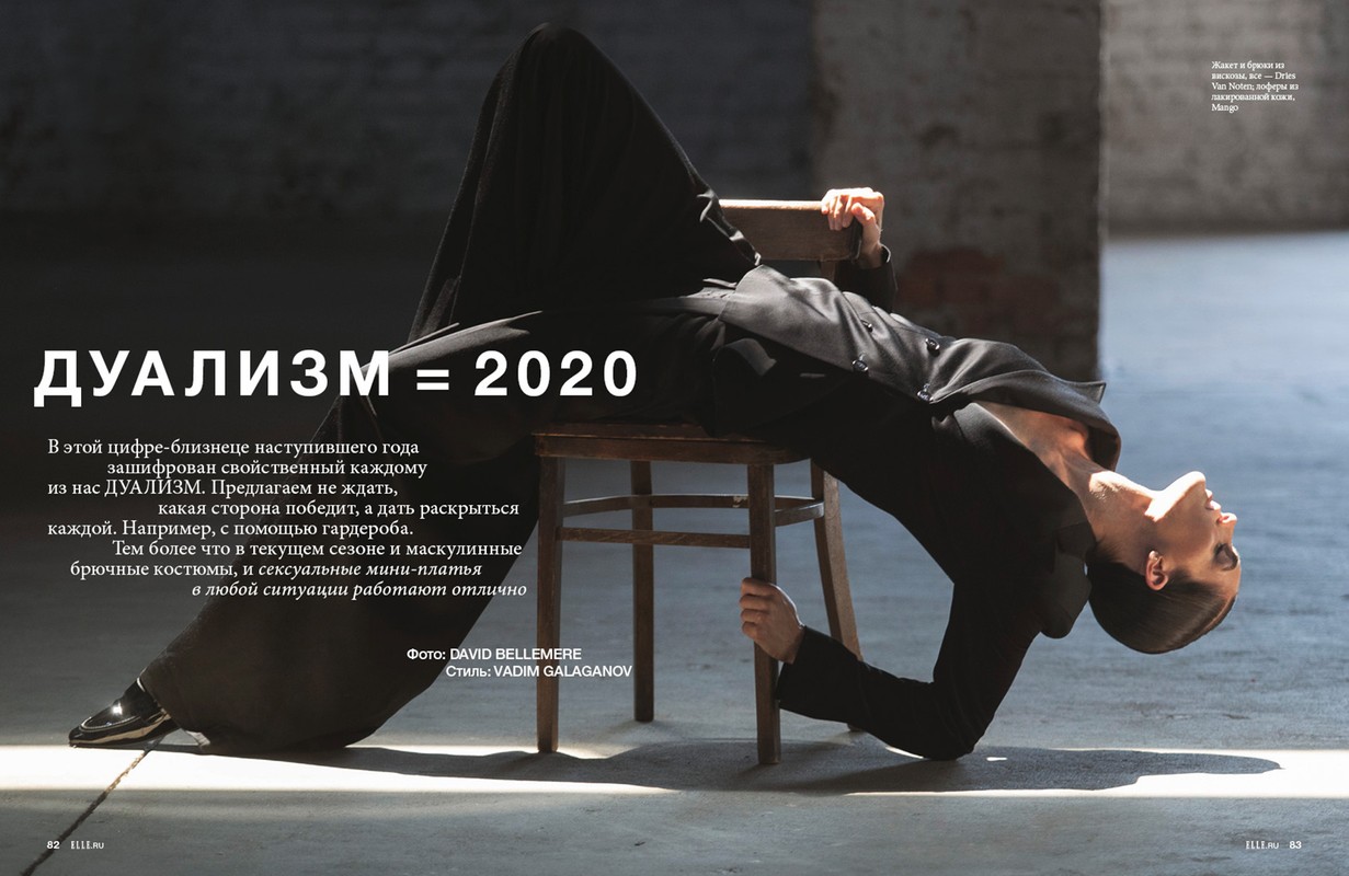 Fashion editorial shot of a bald woman wearing a black suit, leaning backwards in a chair in front of a weathered wall.