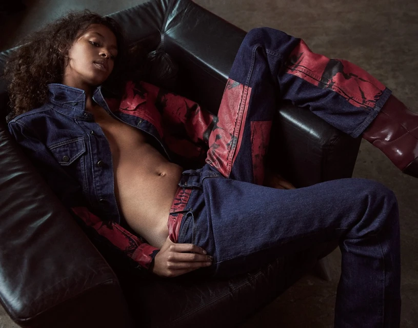 Fashion portrait of a Black woman reclining in a chair, wearing a denim jacket, jeans with leather panels and shiny boots.