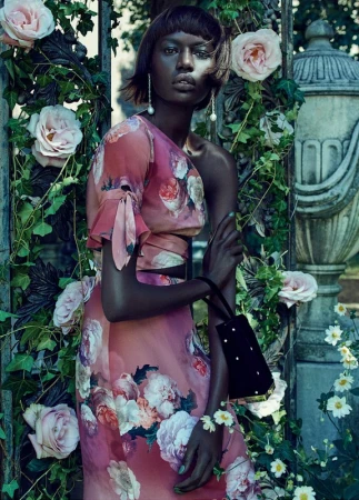 A dark-skinned woman in a floral dress poses in a garden setting with light pink roses and an ornate stone column behind a gate.