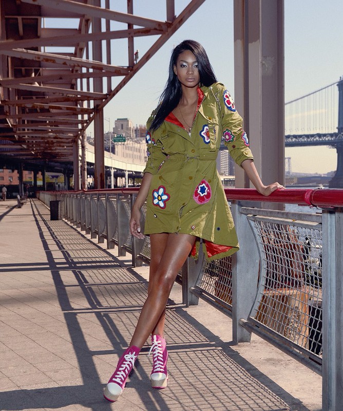 Stylish woman poses in a unique flower-decorated dress and pink sneakers, standing on a bridge with a city and bridge backdrop in daylight.