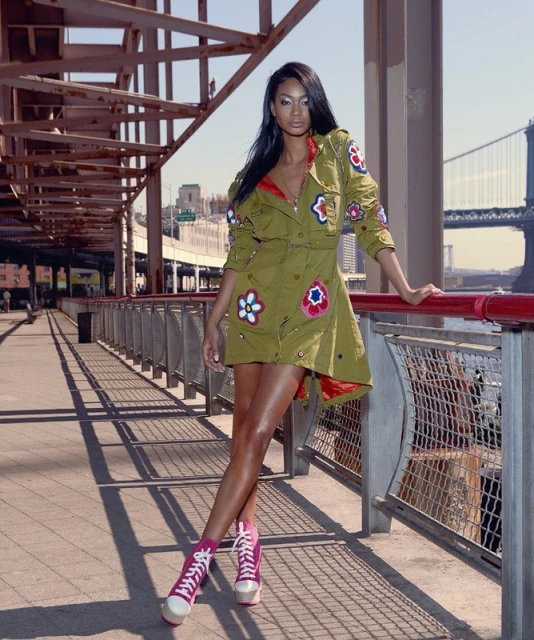Stylish woman poses in a unique flower-decorated dress and pink sneakers, standing on a bridge with a city and bridge backdrop in daylight.