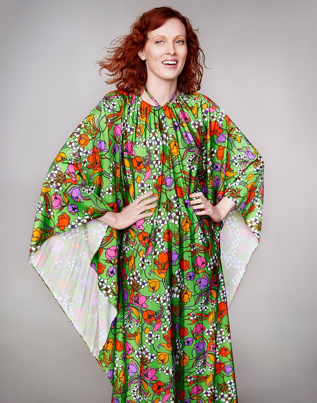 A smiling woman with red curly hair stands with hands on her hips, wearing a green and multi-colored floral print kaftan in a studio.
