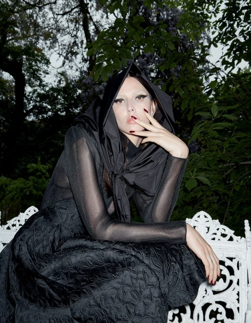 A woman in a black hooded dress poses outdoors with a hand near her face, set against a backdrop of green foliage and a white bench.