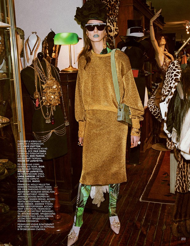 Editorial fashion shot of a female model in a gold knit dress and green leggings in a cluttered vintage clothing store, with accessories and props.