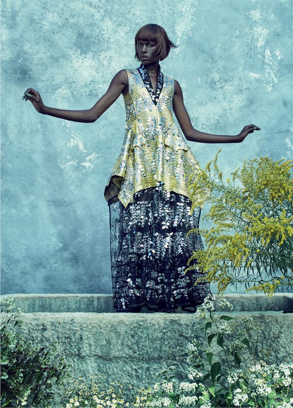 An adult woman with short brown hair poses with arms outstretched on a stone step in a flowing, glittering dress in an outdoor setting.