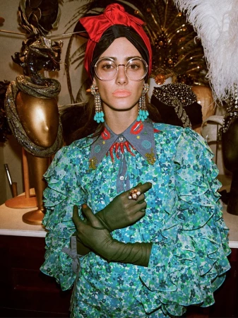 Woman in a patterned dress and red headwrap poses in front of mannequins with decorative hats, featuring turquoise earrings.