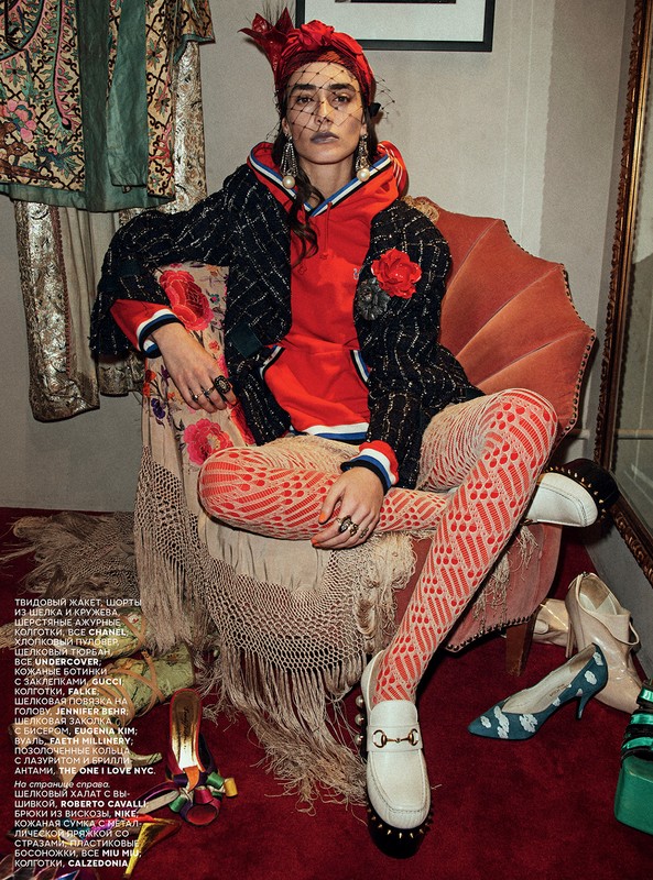 Editorial fashion shot of a woman in a red hooded top, patterned leggings, and a black tweed jacket, seated on a vintage red chair.