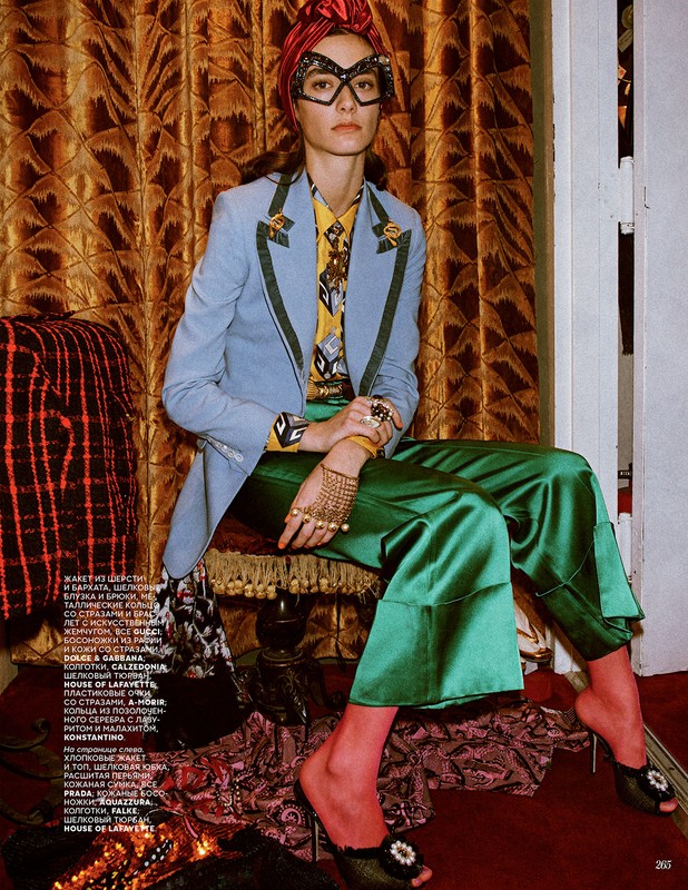 Fashion editorial shot: Woman in turban, glasses, and tailored jacket poses against patterned backdrop, showcasing designer clothing.