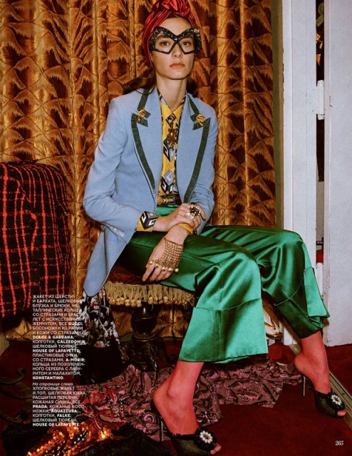 Fashion editorial shot: Woman in turban, glasses, and tailored jacket poses against patterned backdrop, showcasing designer clothing.