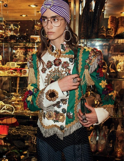 Elegant woman with a turban, eyeglasses, and embroidered jacket indoors amongst glass shelves of accessories and shoes, fashion forward.