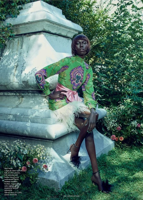 Dark-skinned woman in green patterned dress, sitting on a stone structure in a garden setting, accessorized with feathered heels.