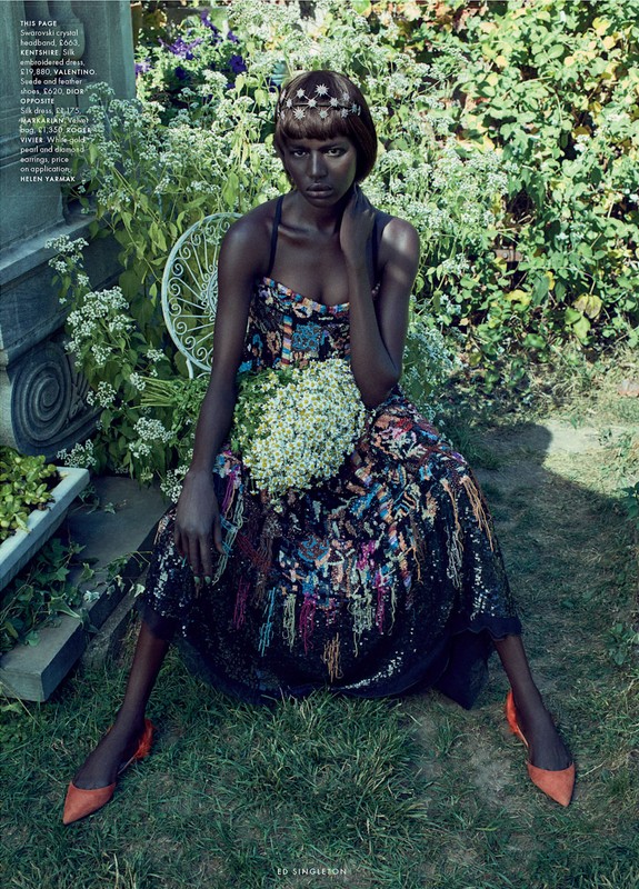A woman in an embellished dress sits in a garden, holding a bouquet, her ensemble completed by orange shoes and a jeweled headband.