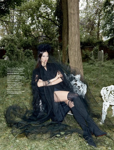 Fashion editorial shot of a woman in black haute couture dress and thigh high boots posing in a graveyard setting with trees.