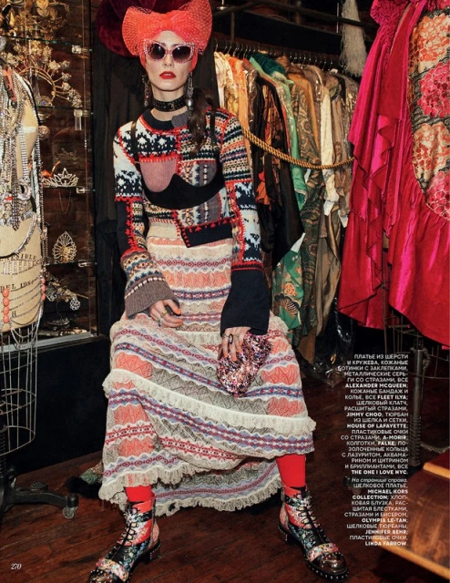 A fashionable adult female sits in a textured sweater, orange skirt, and boots in a vintage clothing shop. She wears round pink sunglasses.