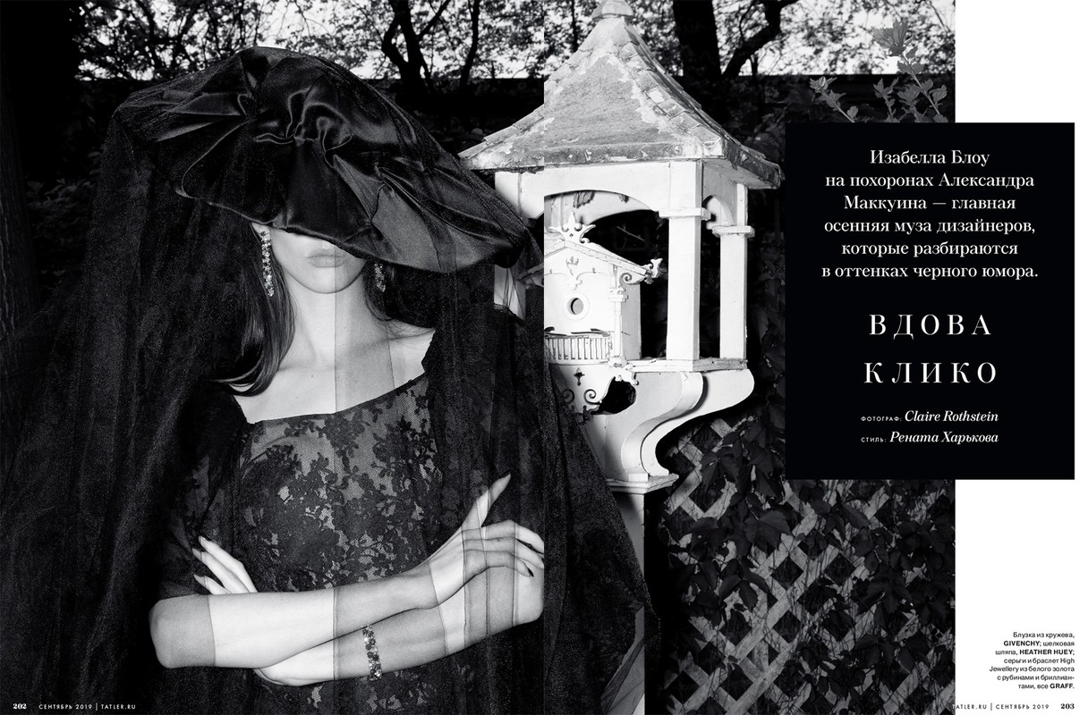 Fashion editorial shot of a woman wearing a black lace dress, a large hat, and a veil, standing outdoors near a decorative birdhouse.
