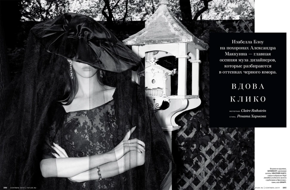 Fashion editorial shot of a woman wearing a black lace dress, a large hat, and a veil, standing outdoors near a decorative birdhouse.