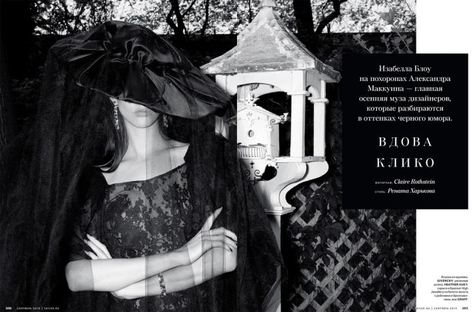 Fashion editorial shot of a woman wearing a black lace dress, a large hat, and a veil, standing outdoors near a decorative birdhouse.