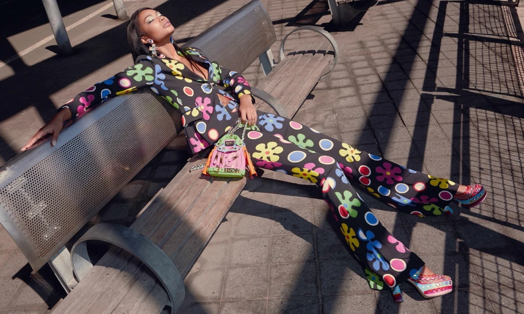 Woman in a colorful flower-print suit poses on a bench outdoors, complemented by a Moschino backpack and sunlight casting shadows.