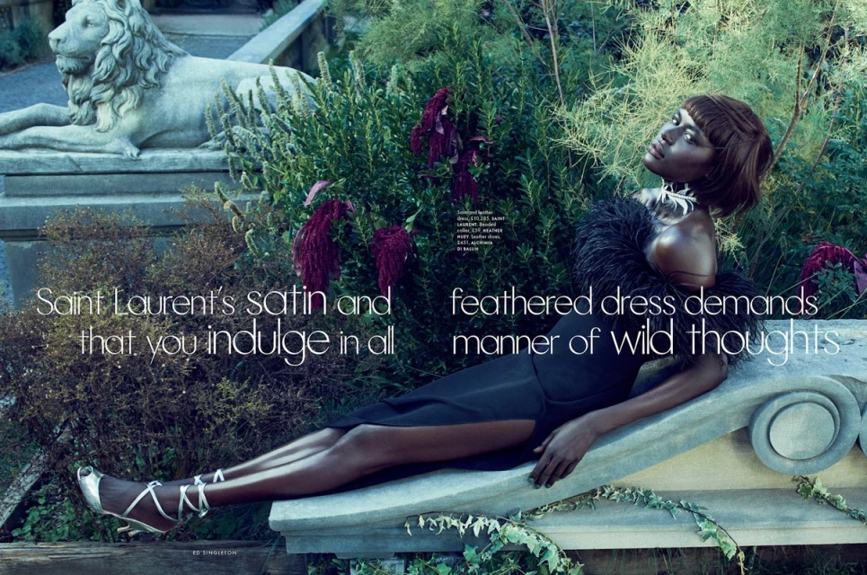 Elegant dark-skinned woman in feathered dress reclines on stone structure surrounded by lush garden foliage and a stone lion statue.