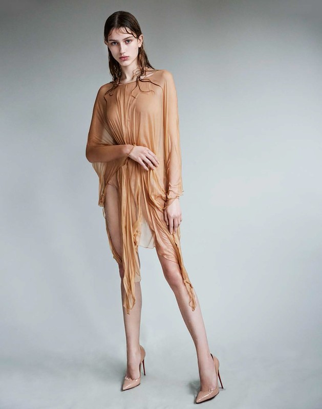 Model with wet hair wearing a sheer draped camel dress and nude heels against a grey studio backdrop