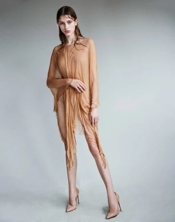 Model with wet hair wearing a sheer draped camel dress and nude heels against a grey studio backdrop