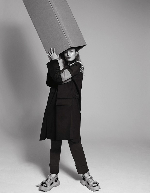 Monochrome fashion shot of a young woman with a cardboard box on her head, wearing an oversized coat and chunky sandals in studio.
