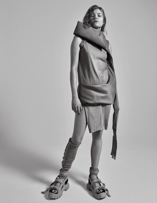 Studio shot of a young woman wearing an avant-garde sleeveless top, skirt and sandal-boots posing in front of a neutral backdrop.
